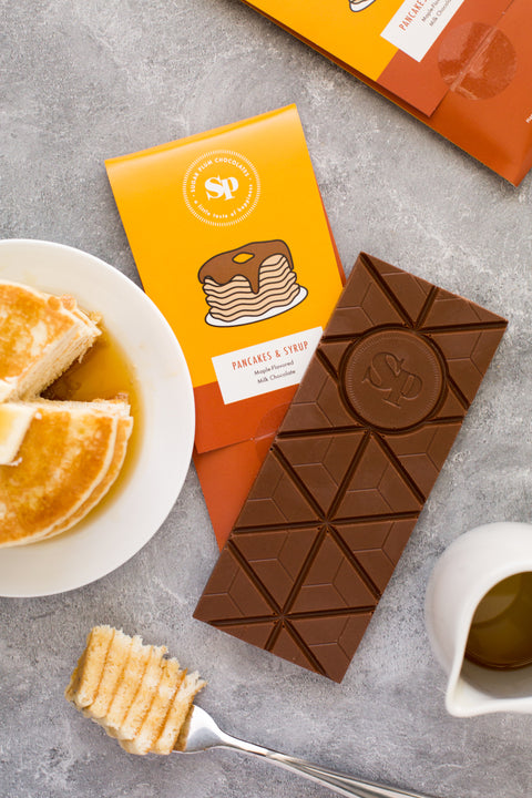 Nostalgic Pancakes & Syrup Chocolate Bar with packaging, next to a plate of pancakes and syrup on a grey surface.