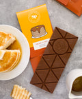 Nostalgic Pancakes & Syrup Chocolate Bar with packaging, next to a plate of pancakes and syrup on a grey surface.