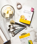Butter seasonings and recipe book with measuring spoons on marble surface, featuring Lemon Garlic spice pack for cooking and grilling.
