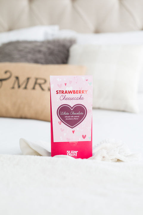 Strawberry Cheesecake White Chocolate Bar in pink packaging on a bed, perfect gourmet dessert gift.