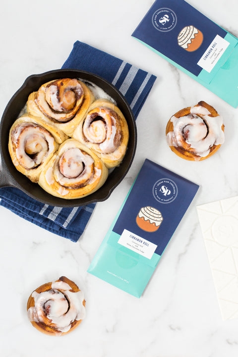 Cinnamon rolls in a skillet with Cinnamon Roll White Chocolate Bars and individual sticky buns on a marble countertop.