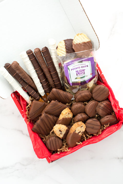 Gourmet chocolate gift box with chocolate-covered pretzels, potato chips, cookies, and assorted treats on a red tissue layer.