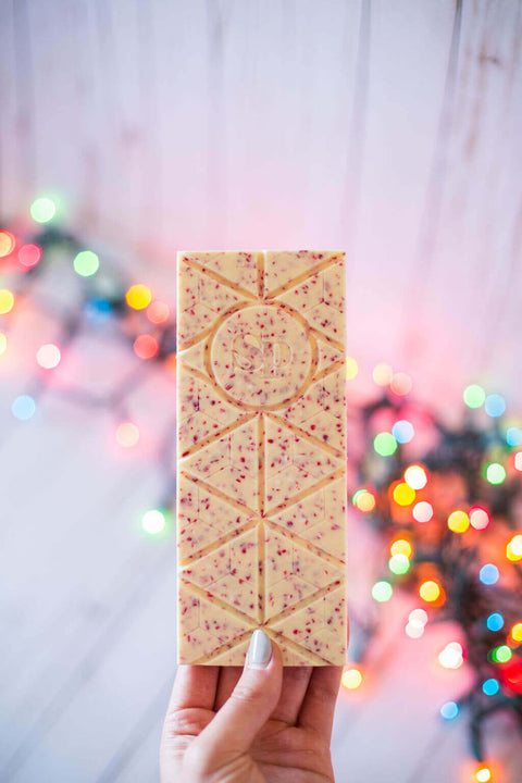 Hand holding a Candy Cane White Chocolate Bar with festive lights in the background, showcasing holiday-themed packaging.