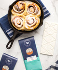 Cinnamon rolls in a skillet with chocolate bars and a checkered napkin on a marble surface.