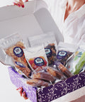Woman holding Sugar Plum Comfort Box filled with 15 gourmet snacks in a purple gift box, perfect for sympathy or get-well gifts.