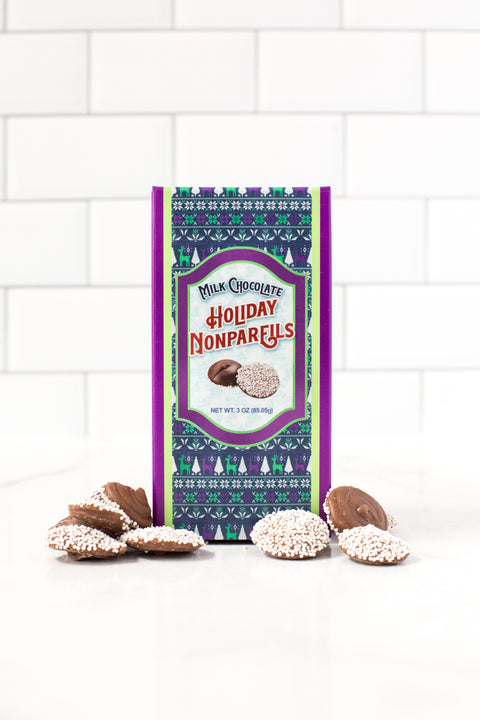 Festive box of milk chocolate holiday nonpareils with scattered peppermint snowflake chocolates on a white tiled background.
