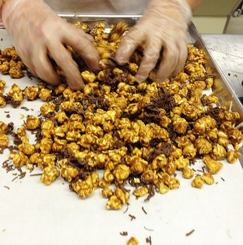 Hands mixing gourmet caramel popcorn with milk chocolate on a tray.