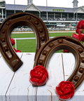 Chocolate horseshoe with red rose, Kentucky Derby theme, on white wooden table in front of racecourse backdrop with roses.