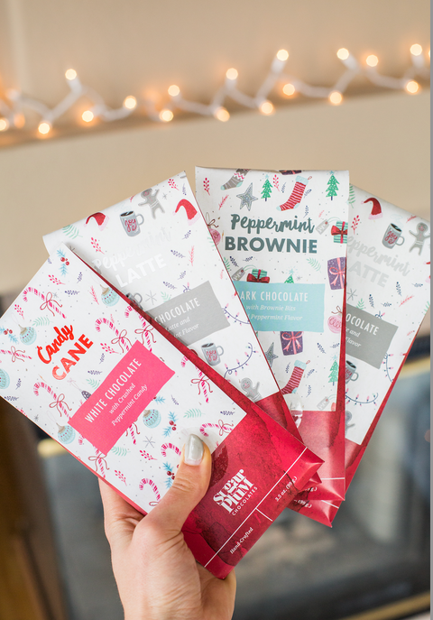 Hand holding Sugar Plum's Peppermint Chocolate Bar Holiday Collection with festive flavors in front of holiday lights.